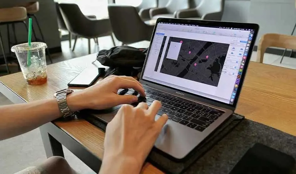 Hands work on an Apple MacBook in a café, map pins on screen, with a phone, bag, and a Starbucks iced drink on the wooden table.