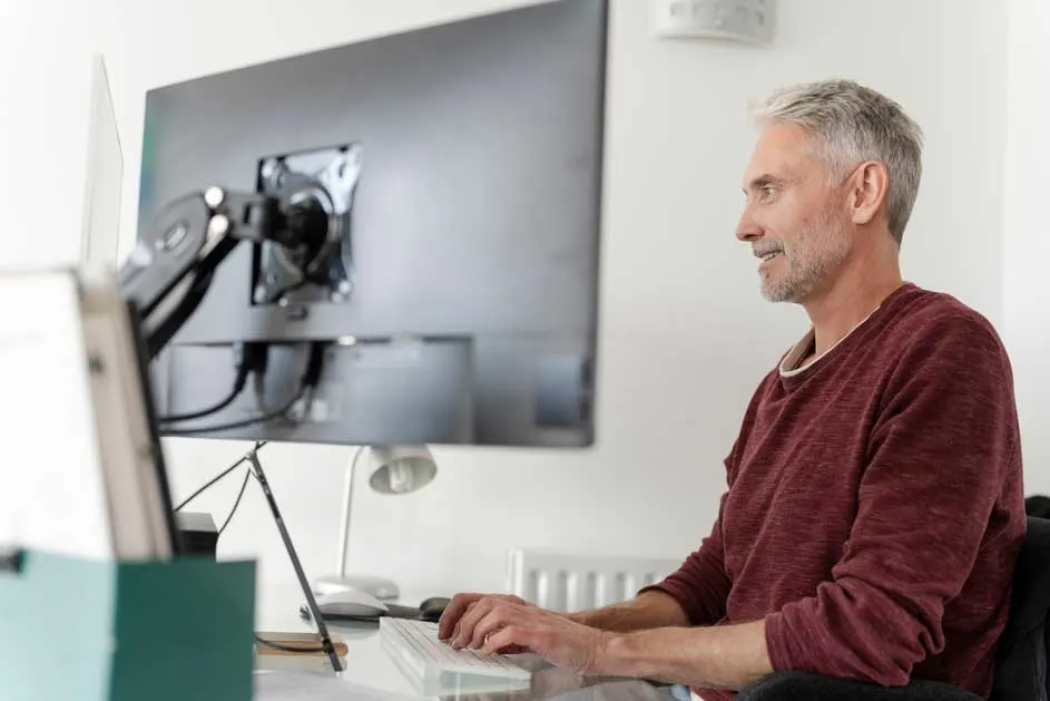 Un grand écran d'ordinateur est monté sur un bureau blanc, avec un clavier en avant-plan. Un support maintient l'écran.