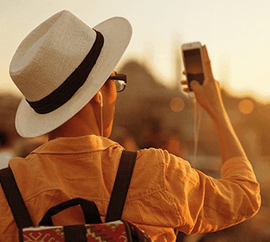 Un homme avec un chapeau beige et un sac à dos prend une photo avec son téléphone devant un paysage urbain au coucher du soleil.