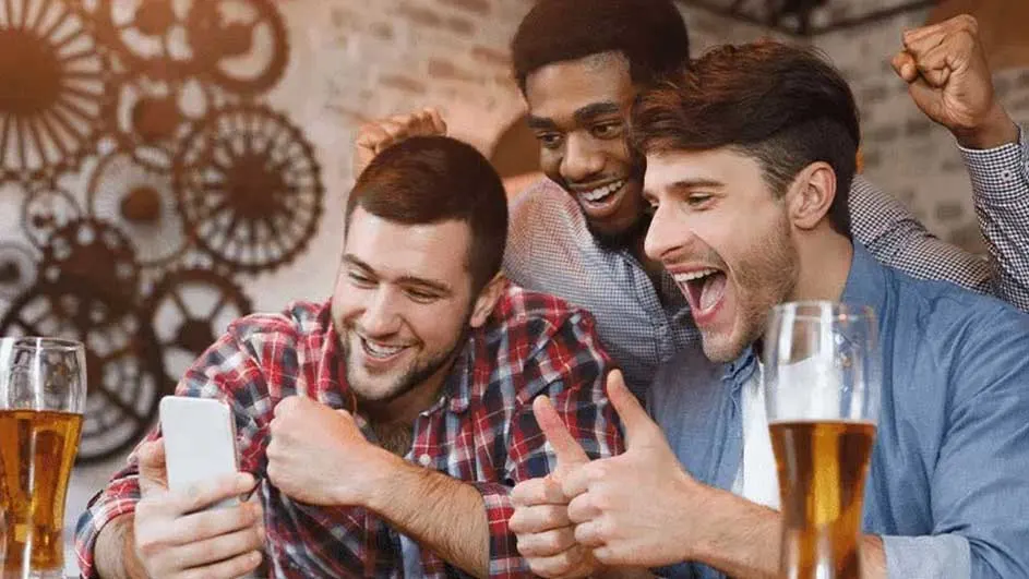 Trois hommes sourient vers un téléphone avec des bières à l'avant et un mur avec des décorations d'engrenages à l'arrière-plan.