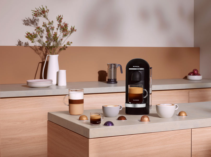 Een keukenopstelling met een Nespresso apparaat, diverse koffiekopjes en capsules op een lichtgrijs aanrecht.