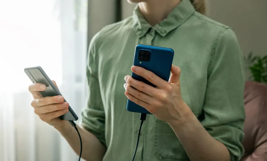 Een persoon houdt twee smartphones vast, een blauwe aan de rechterkant en een grijze aan de linkerkant, beide met een kabel.