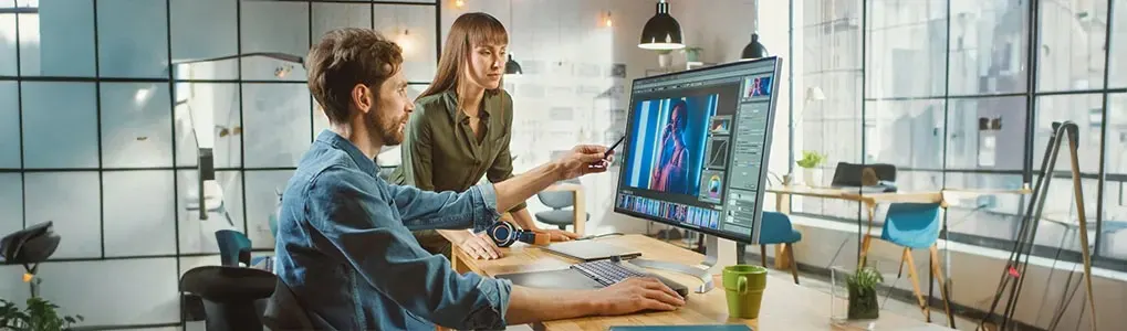 Deux personnes travaillent dans un bureau moderne sur la retouche d’image, devant un grand écran, avec vitres, plantes et matériel.