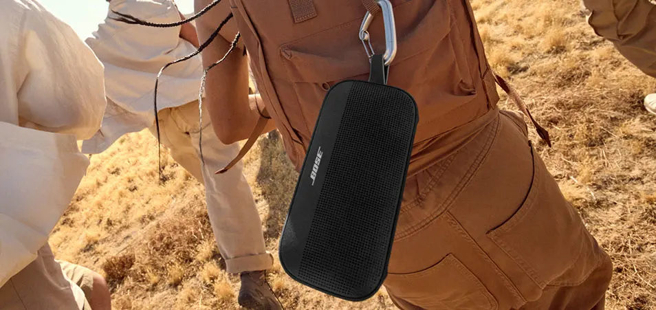 Een zwarte Bose-luidspreker hangt aan een zilveren karabijnhaak aan een bruine rugzak, tegen een achtergrond van droog gras.
