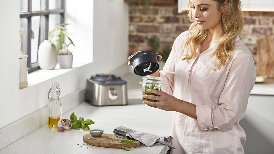 Een keuken met een blender, kruiden en een snijplank op een wit aanrechtblad. Een raam en stenen muur zijn zichtbaar.