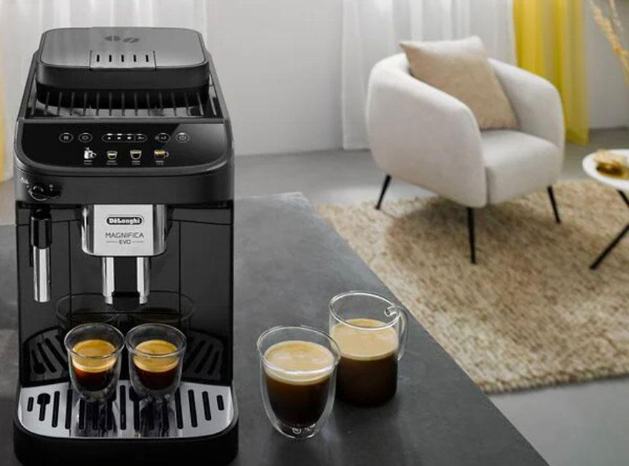 Machine à café noire avec deux tasses remplies, sur une table grise. Un fauteuil beige et un tapis sont visibles en arrière-plan.