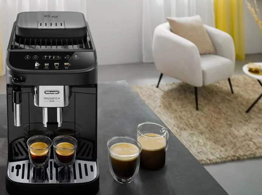 Machine à café noire avec deux tasses remplies, sur une table grise. Un fauteuil beige et un tapis sont visibles en arrière-plan.