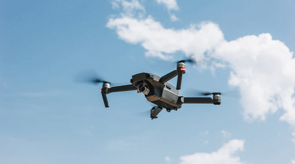 Een grijze drone vliegt tegen een blauwe lucht met witte wolken op de achtergrond. De propellers draaien.