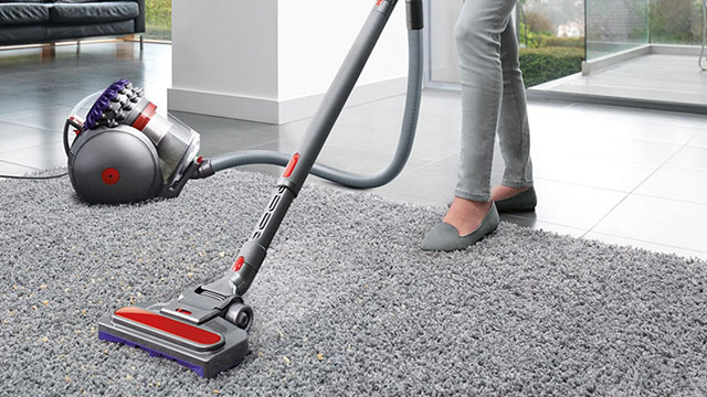 Aspirateur gris avec tube sur tapis gris. Canapé noir et sol blanc en arrière-plan.