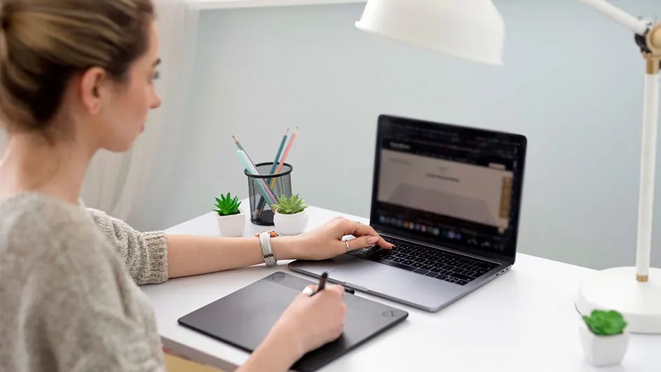 Personne travaille sur un MacBook Apple avec une tablette graphique, bureau blanc, plantes, lampe et pots à crayons ; logo Apple visible.