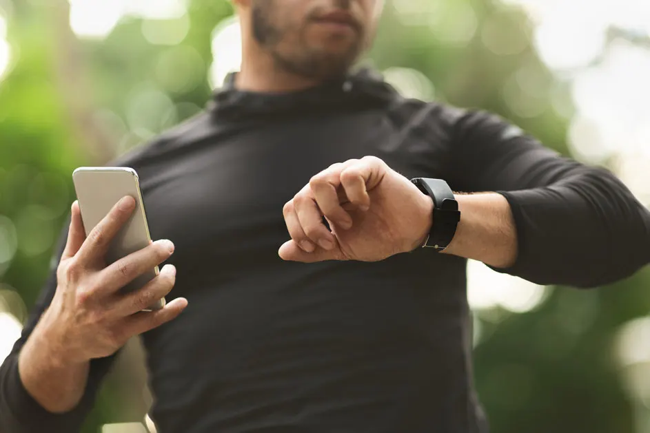 Een persoon in een zwart shirt controleert een zwart horloge en houdt een smartphone vast tegen een groene achtergrond.