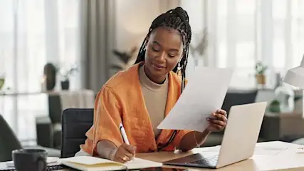 Een bureau met een laptop, notitieboek en papieren. Er staat een pen klaar, en er is natuurlijk licht.