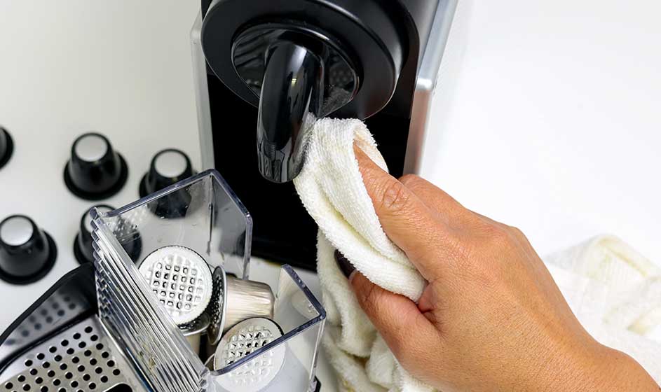 Een hand maakt de tuit van een koffiemachine schoon met een doek, naast een uitneembaar bakje en losse onderdelen op het aanrecht.