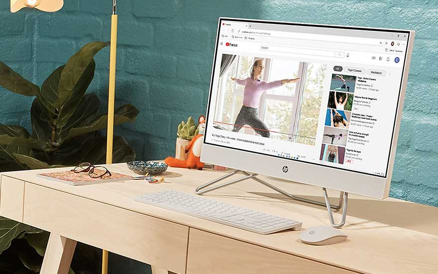 Bureau beige avec ordinateur affichant une vidéo, clavier blanc et souris. Plante verte et lampe jaune en arrière-plan.