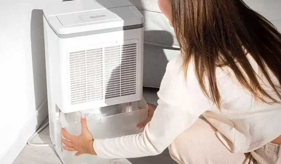 Un déshumidificateur blanc avec grille est sur un sol clair. Une personne retire le bac à eau transparent.