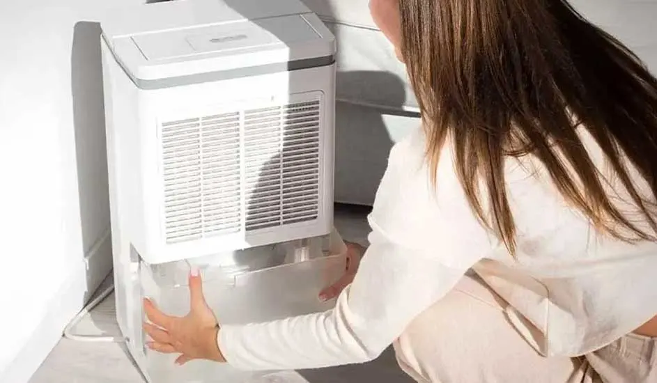 Un déshumidificateur blanc avec grille est sur un sol clair. Une personne retire le bac à eau transparent.