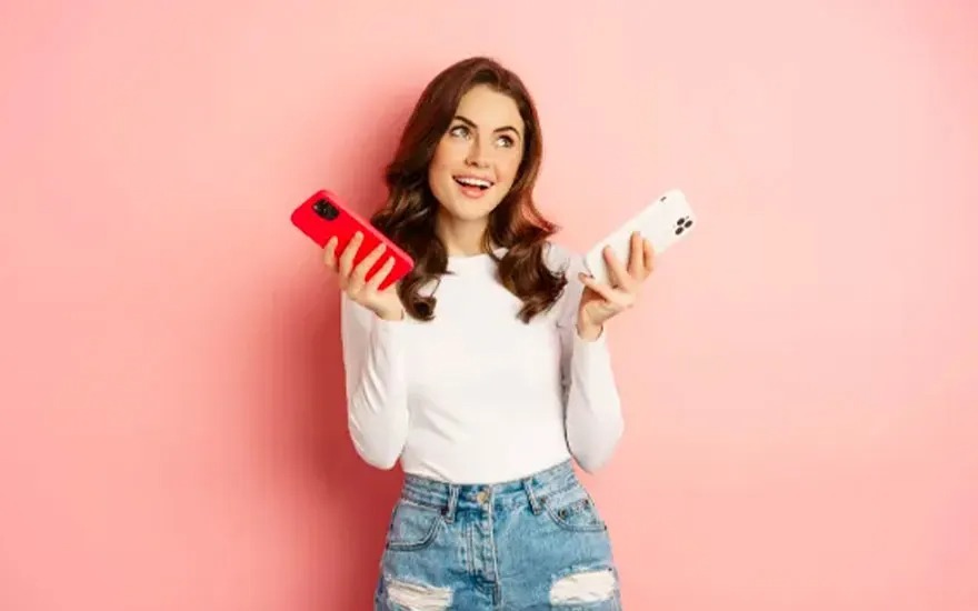 A person holds a red and white phone against a pink background. The person wears a white shirt and blue jeans.