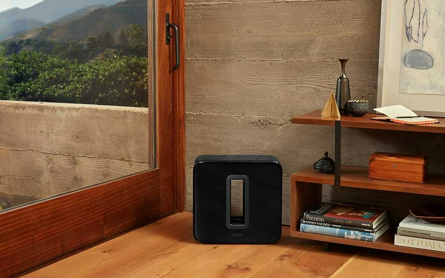 Een zwarte subwoofer staat op een houten vloer naast een raam met uitzicht op bergen en een houten rek met boeken en decoraties.