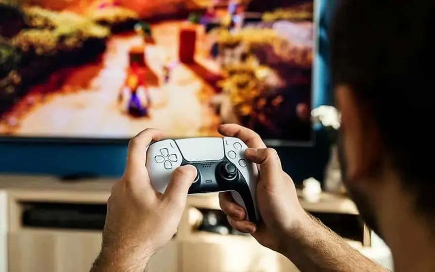 A person is playing a colourful game on a PS5 using a white DualSense controller, facing a large television in a living room.