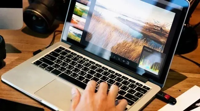 A person edits a vivid landscape photo on a silver laptop, while a camera and small accessories rest beside it on the wooden desk.