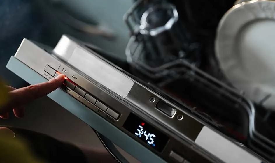 A finger presses the buttons on a dishwasher. The digital display shows 35 degrees, while plates and cutlery are visible inside the rack.