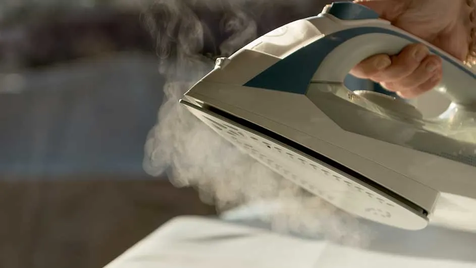Een stoomstrijkijzer met blauwe accenten wordt gebruikt; stoom komt uit de zoolplaat op een wit oppervlak.