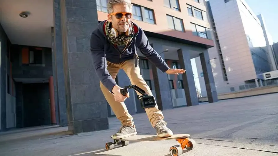 A skateboard on pavement with buildings in the background. It has orange wheels. A camera is attached to the skateboard.