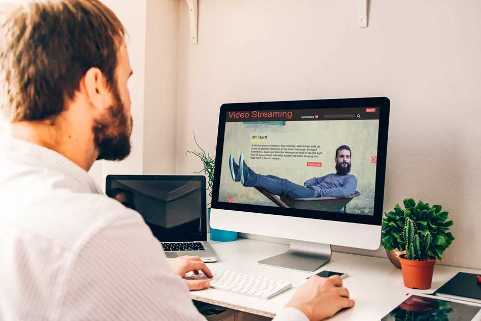 Un homme utilise un ordinateur pour regarder des vidéos en streaming sur un bureau blanc avec des plantes.
