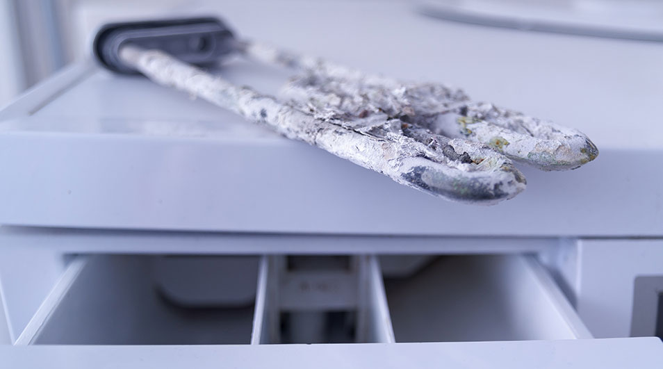 Twee witte, kalkaanslag bedekte verwarmingselementen liggen op een open wasmachinecompartiment.