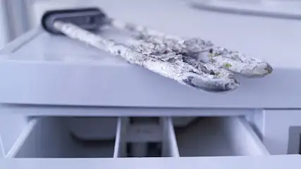 Twee witte, kalkaanslag bedekte verwarmingselementen liggen op een open wasmachinecompartiment.