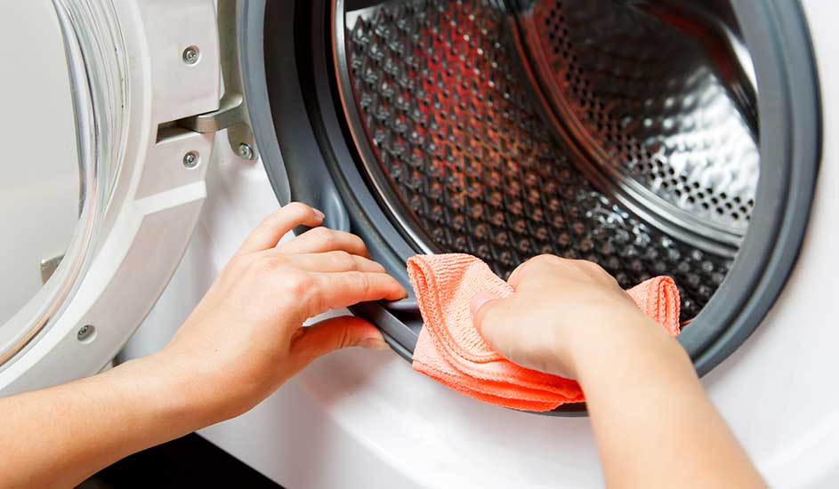 Une main essuie le joint gris d'un lave-linge blanc ouvert avec un chiffon orange. Le tambour en métal est visible.