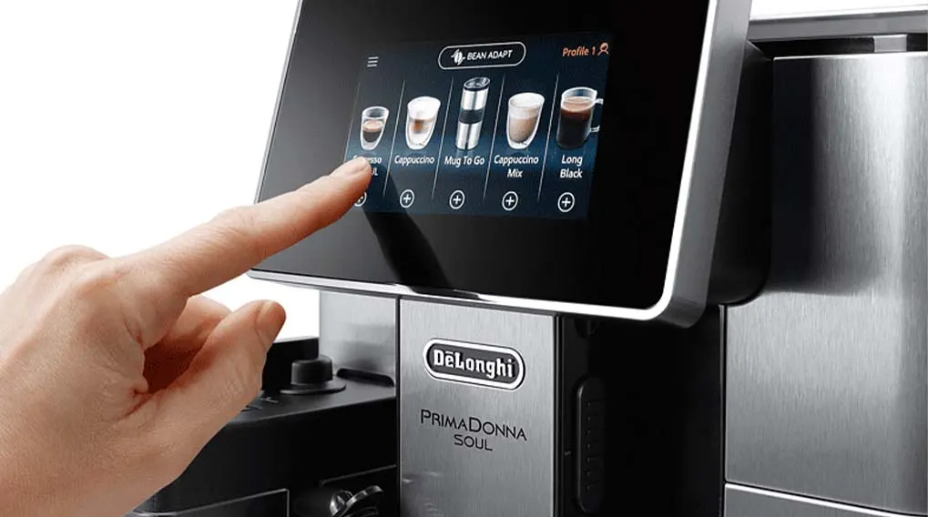 Een zilverkleurige DeLonghi Primadonna Soul koffiemachine met een touchscreen waarop koffieopties staan afgebeeld. Een vinger selecteert een drankje.