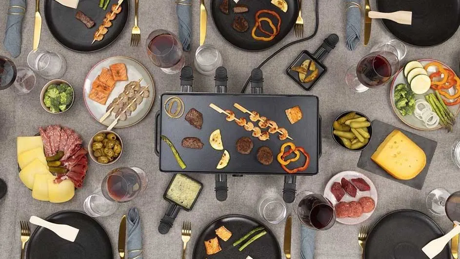 Vue aérienne d'une table de raclette avec grill, plats préparés, fromage, légumes, charcuterie et verres de vin.