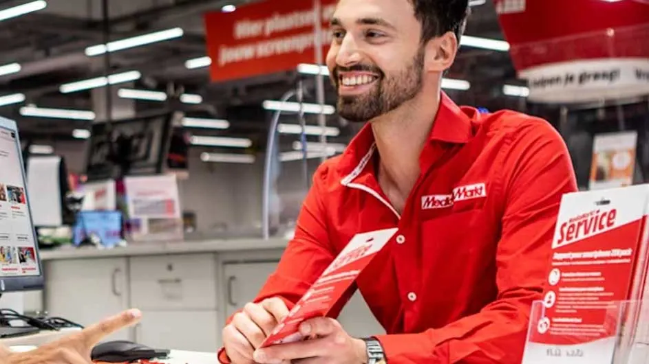 Een man in een rood Media Markt-shirt staat achter een balie met een service-folder. Op de achtergrond hangen reclameborden.