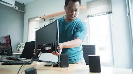 Bureau avec ordinateur, clavier, souris, haut-parleurs noirs. Un homme tient un écran. Fenêtre claire en arrière-plan.
