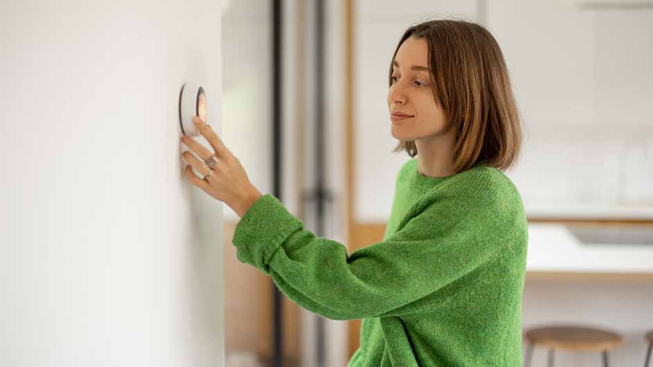 Une main ajuste un thermostat rond blanc sur un mur blanc. Un pull vert est visible. Arrière-plan : mobilier de cuisine clair.