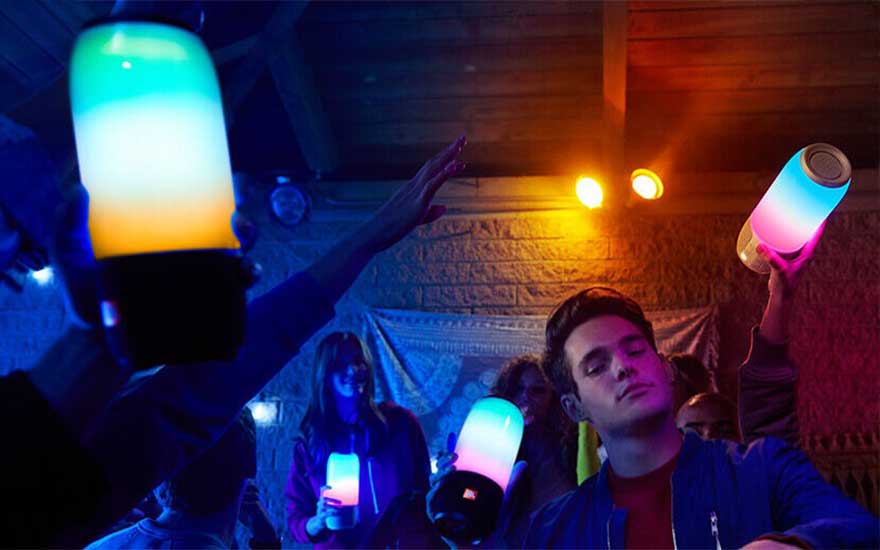 Mensen houden kleurrijke luidsprekers vast in een kamer met blauw en oranje licht. De muur heeft een stenen textuur.