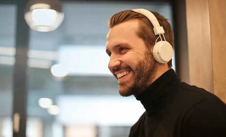 Un homme porte un col roulé noir et un casque blanc. En arrière-plan, on aperçoit une fenêtre et un luminaire.