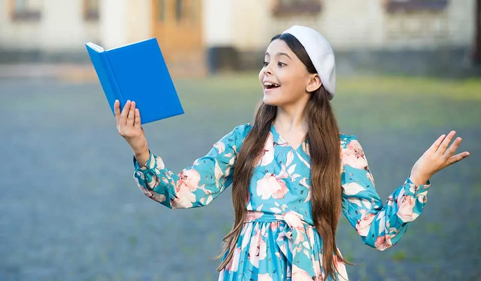 Une personne en robe à fleurs bleu clair tient un livre bleu. Elle porte un béret blanc devant un bâtiment.