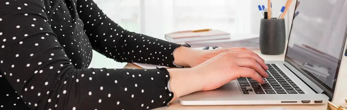 Personne en blouse noire à pois blancs tape sur un MacBook Apple ; carnets et pot à crayons sur le bureau, design Apple reconnaissable.