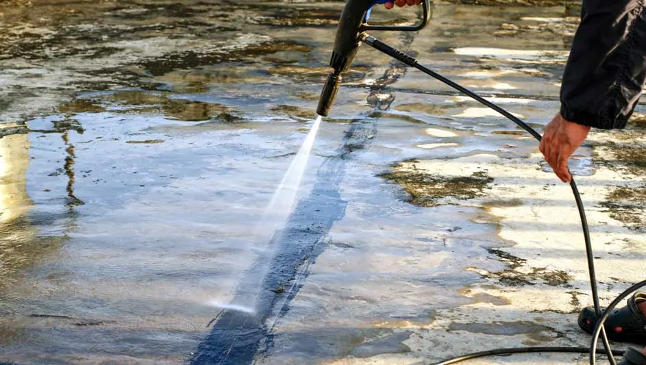 Een hogedrukreiniger spuit water op een betonnen oppervlak, waardoor een schoon pad ontstaat. Het vuile gebied is grijs en bruin.