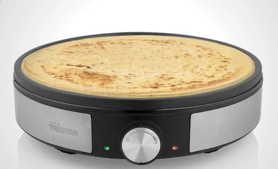 Crêpière électrique noire et argentée avec une crêpe cuite dessus. Une molette de réglage est visible sur le devant.