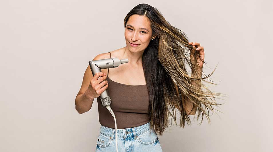 Une femme tient un sèche-cheveux gris, cheveux longs bruns et blonds, débardeur marron, jean bleu clair. Fond uni.