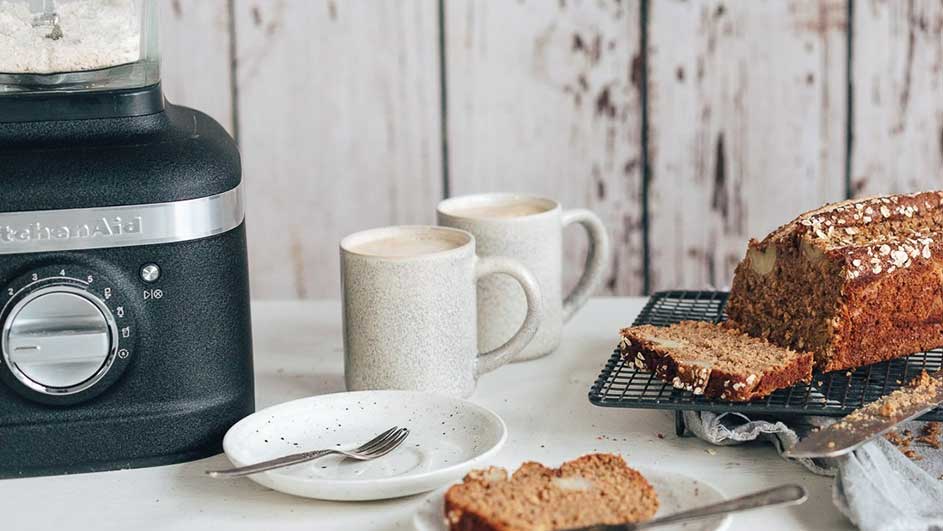 Een zwarte keukenrobot, twee mokken, en een gesneden brood staan op een witte tafel tegen een houten achtergrond.