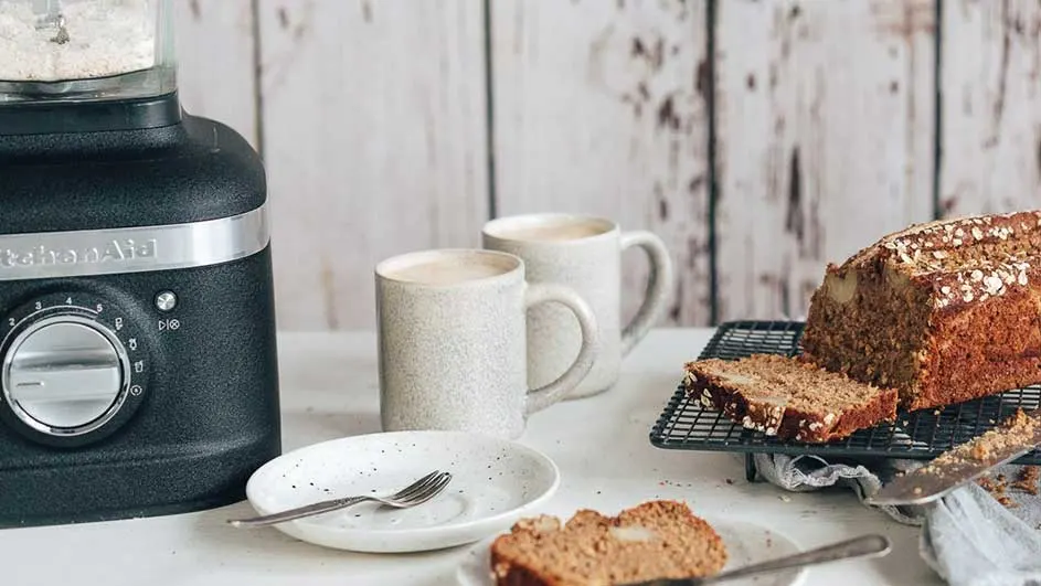 Een zwarte keukenrobot, twee mokken, en een gesneden brood staan op een witte tafel tegen een houten achtergrond.