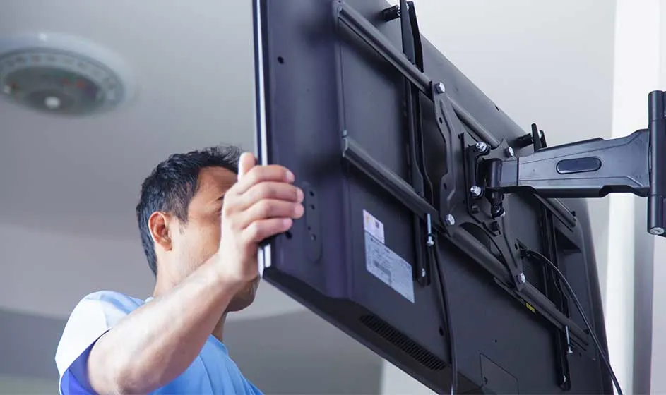 A man is mounting a large television on a black wall bracket. He holds the screen while the metal structure and cables remain visible at the back.