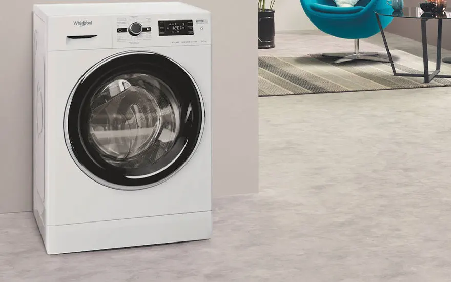 Witte wasmachine met een zwarte deur tegen een lichtgrijze muur. Op de achtergrond een blauwe stoel en een zwart tafeltje.