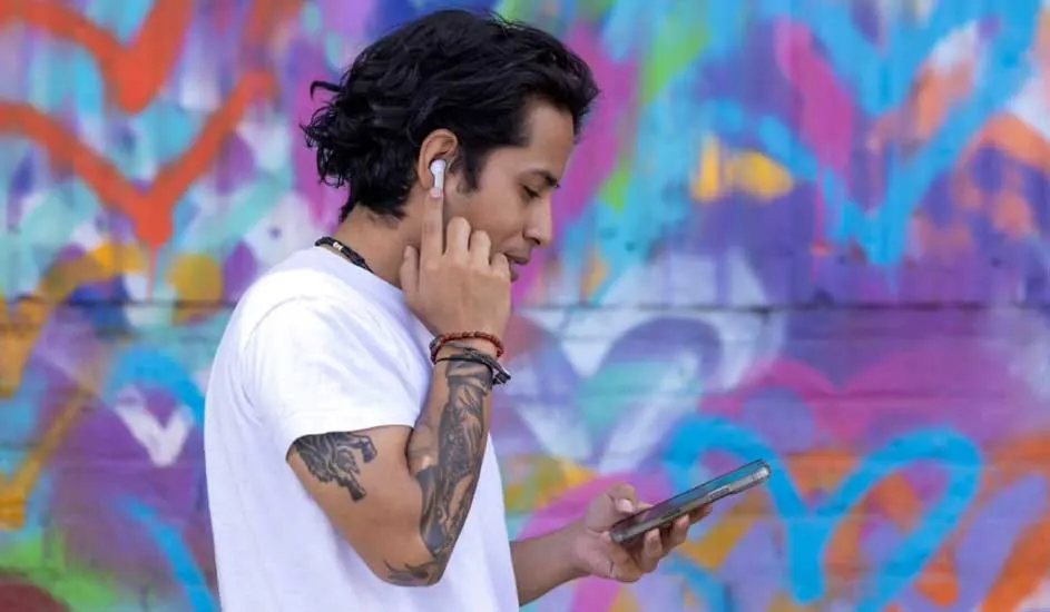 Un homme ajuste son écouteur sans fil, téléphone en main, devant un mur de graffitis colorés.