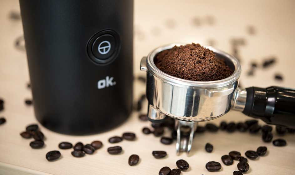 Een zwarte koffiemolen staat naast een metalen filterhouder vol gemalen koffie. Rondom liggen verspreide koffiebonen op een lichte houten tafel.