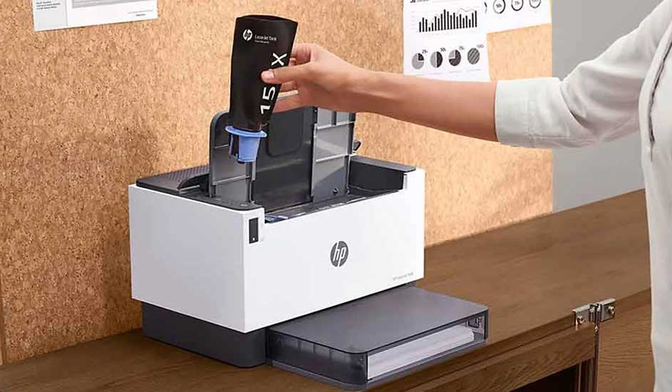 Een persoon plaatst een zwarte HP 15X tonercartridge in een witte HP laserprinter op een houten bureau.